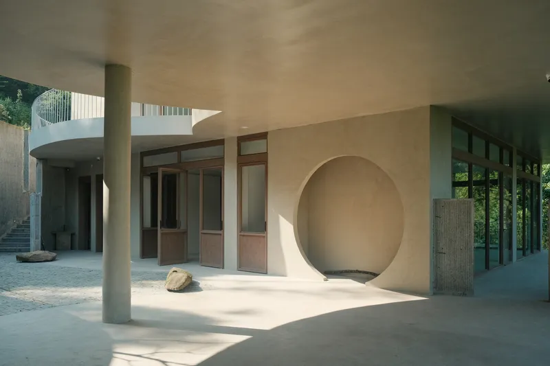 Covered courtyard with cylindrical columns, timber-framed glazing, and circular opening in plaster wall
