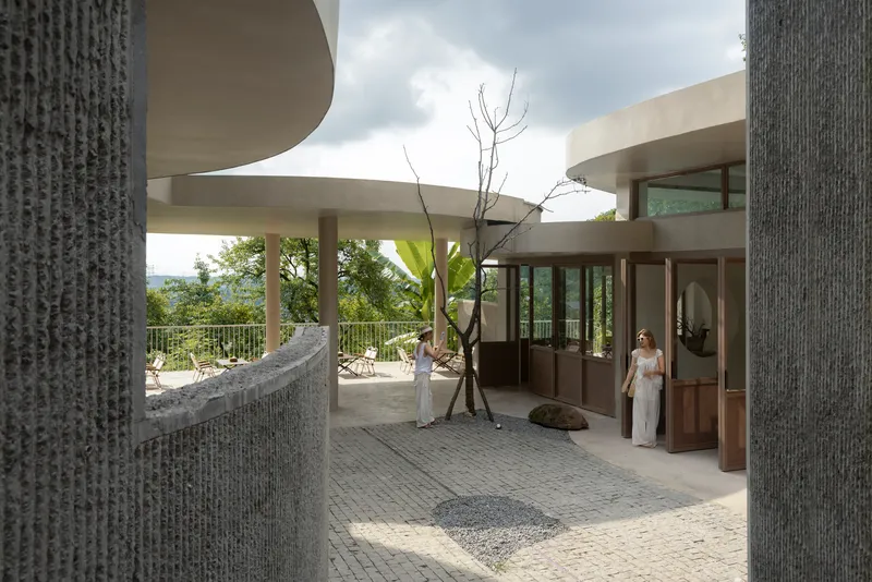 Courtyard terrace with ribbed columns and curved walls beneath overhanging roof lobes under cloudy skies
