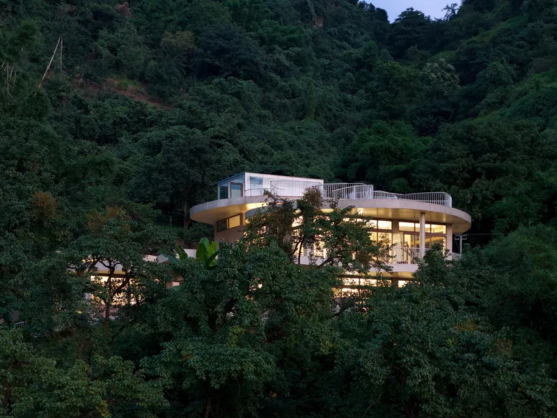 Curved glass facade nestled into dense jungle hillside at dusk with interior lights glowing