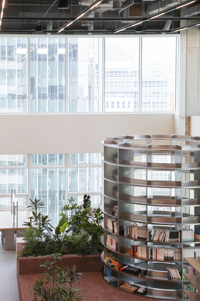 Spiral bookshelf beside planters in a double-height space with exposed ceiling services and curtain wall windows