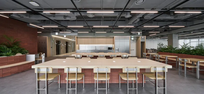 Large communal table with metal stools beneath an exposed structural ceiling grid