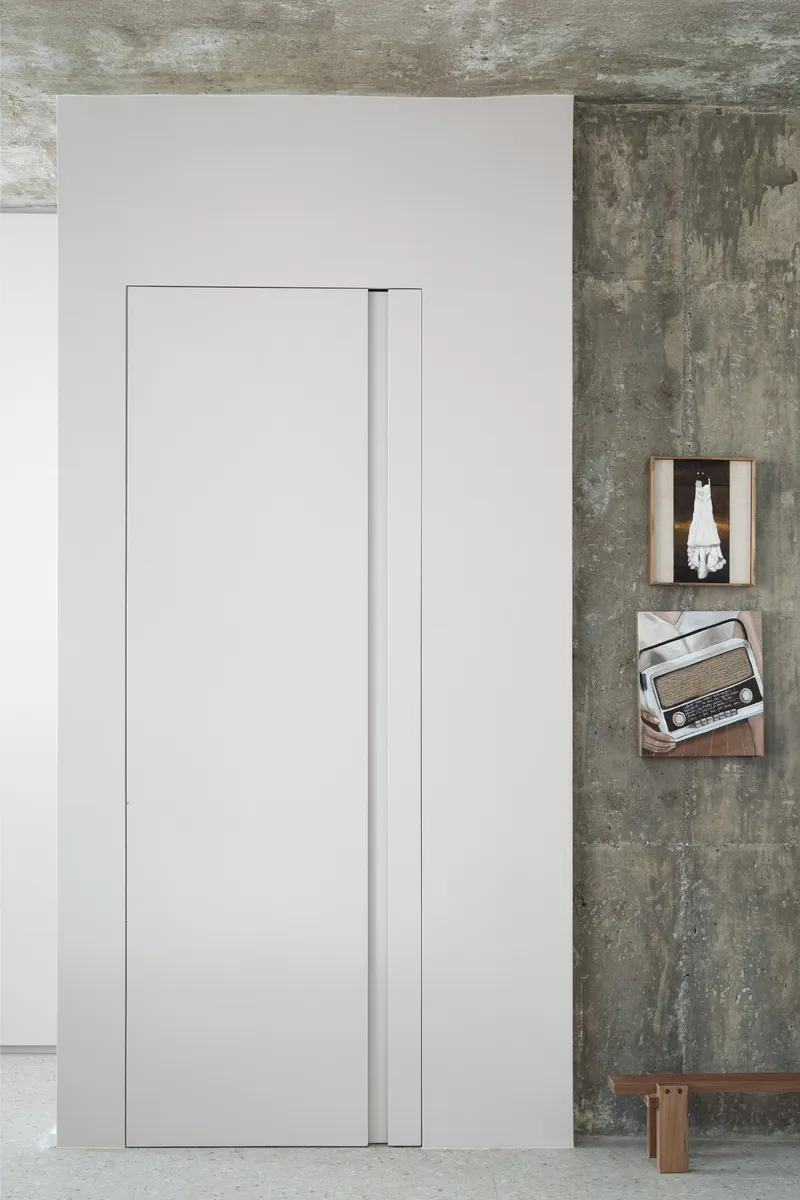 Flush white pocket door beside exposed concrete wall with wooden shelves and a vintage radio