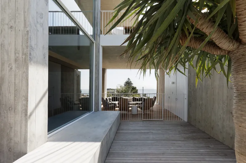 Timber deck passage beneath a concrete soffit with palm fronds overhead and sea view beyond