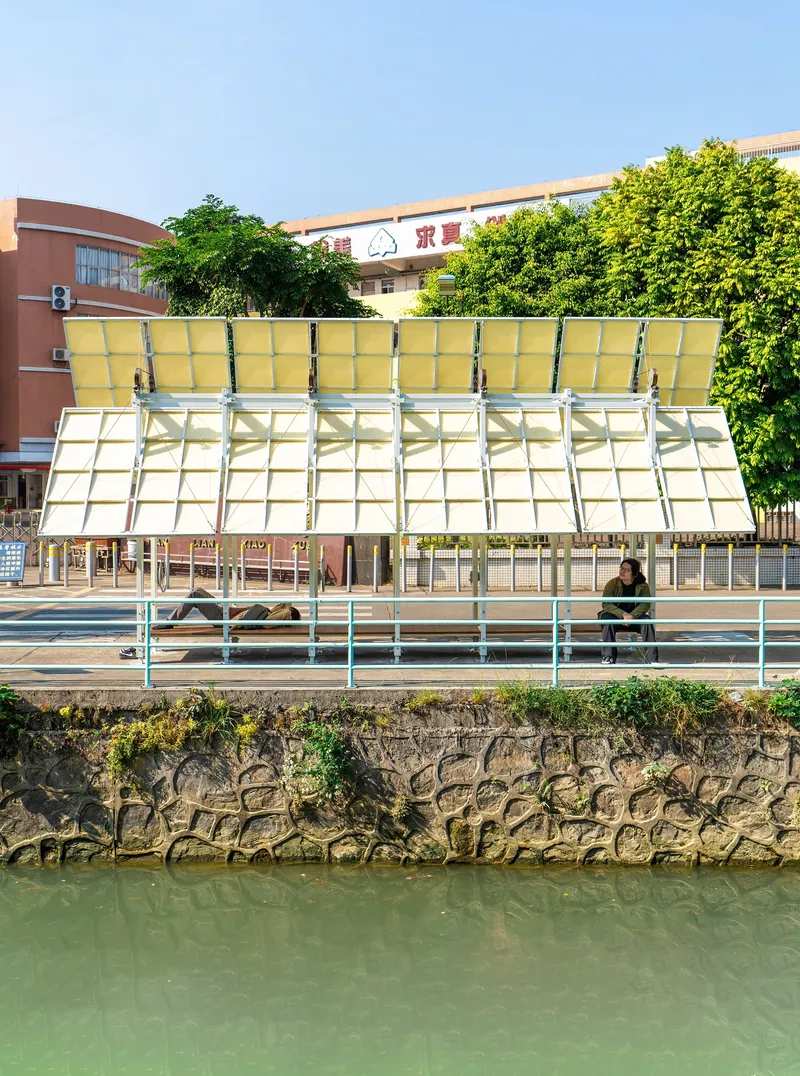 Sloped canopy with translucent yellow panels and steel frame above a stone embankment wall