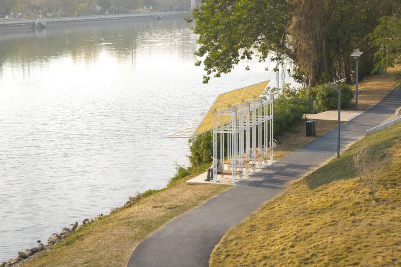 Timber and white steel pavilion with translucent roof panels along a riverside path at sunset