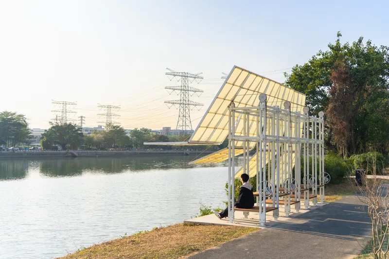 Angled pavilion with white columns and yellow translucent roof beside a lake with power lines beyond