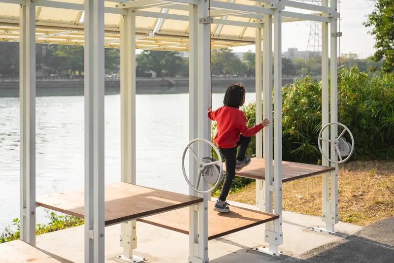 Child in red sweater standing on timber bench inside white steel pavilion overlooking water