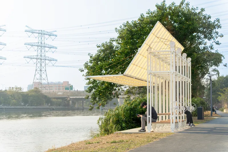 Riverside shelter with angled translucent roof and white vertical slats framed by power lines and trees