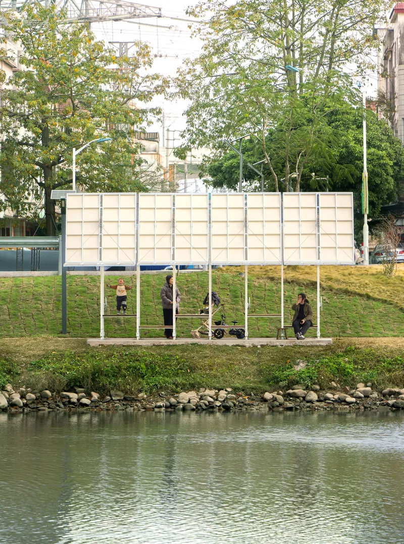 Long pavilion with grid of translucent panels along a canal path with visitors seated beneath
