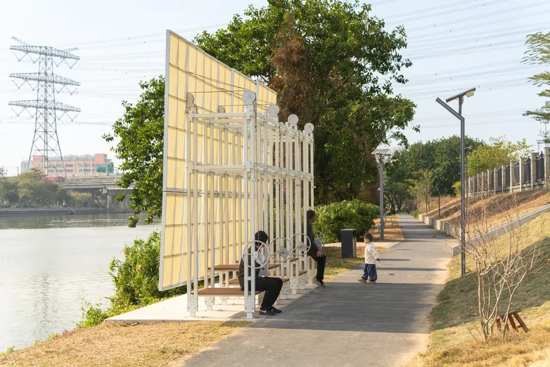 Angled shelter with translucent panels and white framework along a waterside path with pedestrians passing