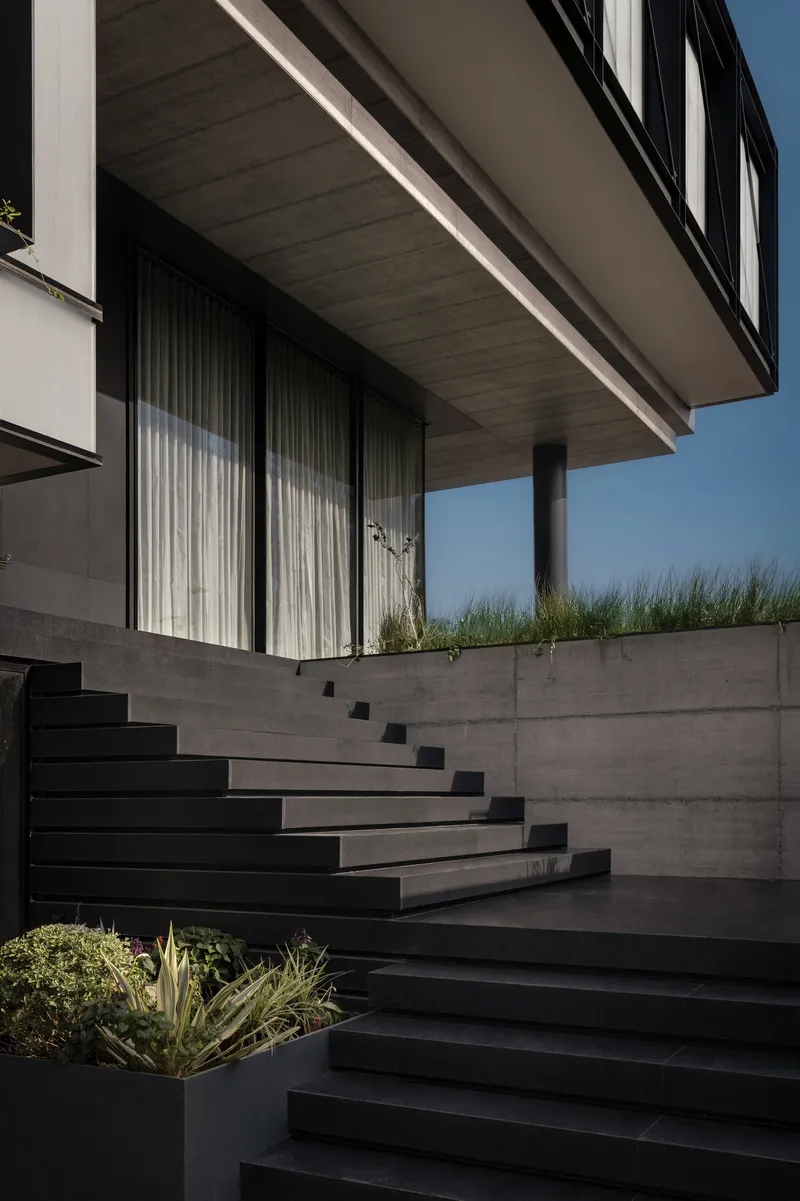 Black steel stair rising along concrete retaining wall beneath cantilevered volume with planted grasses