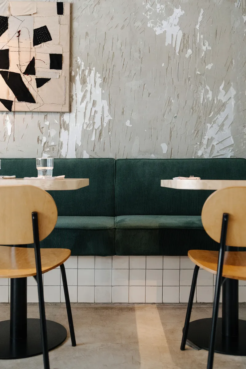 Dining area with green upholstered banquette, round timber tables and weathered plaster wall with peeling paint