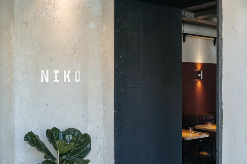 Entrance view with textured white wall, fiddle-leaf fig plant and dark doorway revealing booth seating inside