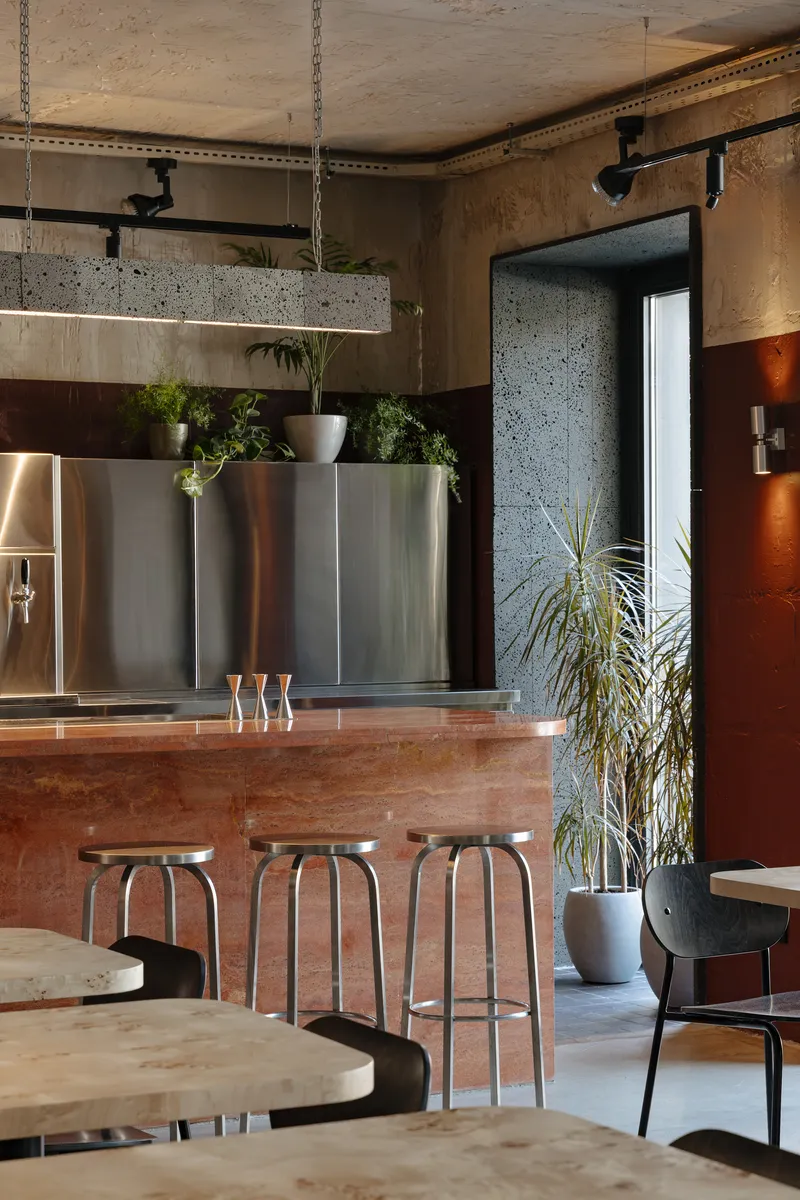 Bar counter with oxidized copper face and metal stools beneath stainless steel back bar and suspended plants