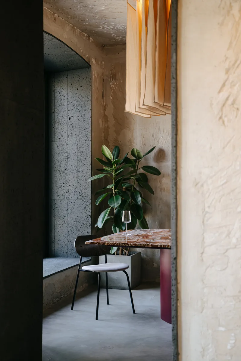 View through a doorway into a dining area with exposed concrete walls and a potted plant