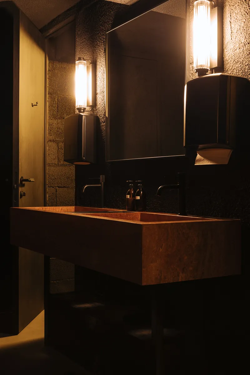 Restroom vanity with copper sink, textured concrete walls and cylindrical wall sconces under low ambient lighting