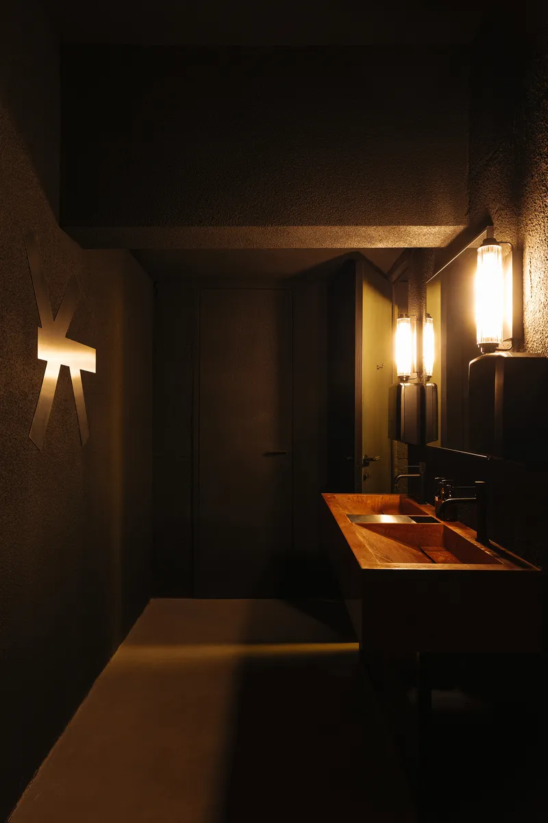 Restroom interior showing copper vanity, concrete ceiling and walls with wall sconces illuminating the space at night