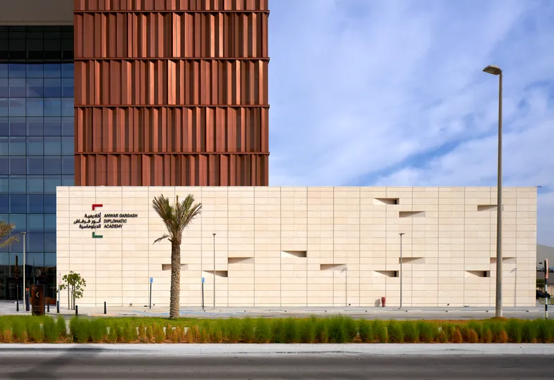 Cream stone facade with recessed window openings beside a copper-clad tower and planted median under blue sky