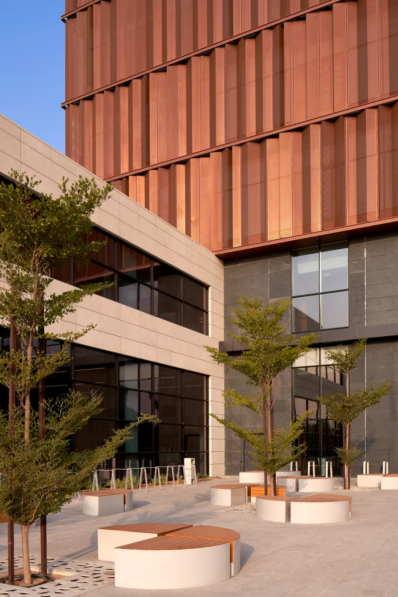 Courtyard with curved stone benches and young trees beneath copper-clad and glass-clad facades in afternoon light
