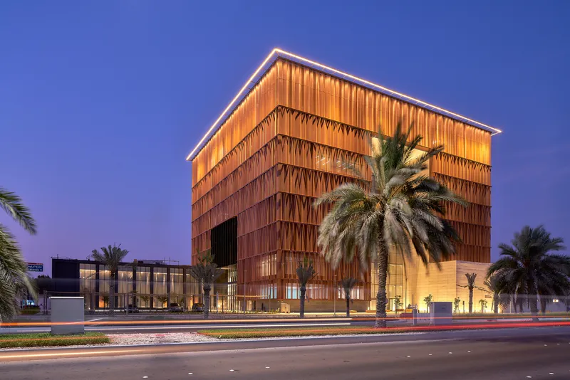 Street view of the copper-screened tower beside date palms with light trails from passing traffic at twilight