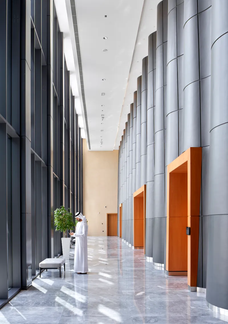 Tall entrance corridor with fluted metal columns and polished marble floor as figure walks through sunlight