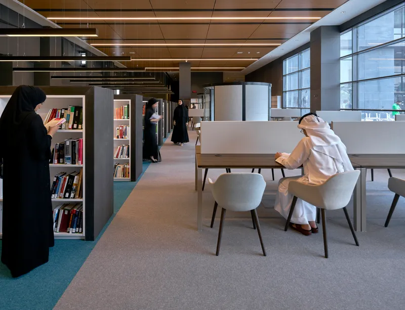 Library reading area with grey shelving and tables where visitors browse books beneath linear ceiling lights