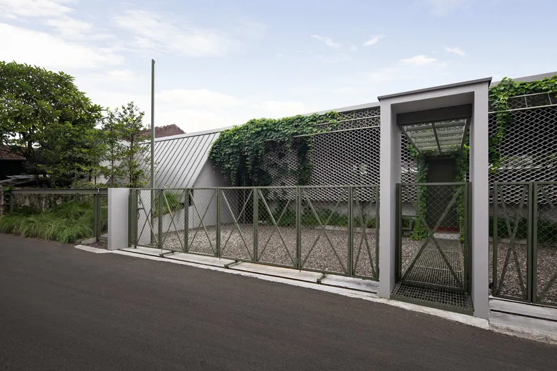 Street view of perforated metal fence with gate opening and climbing vines under a partly cloudy sky