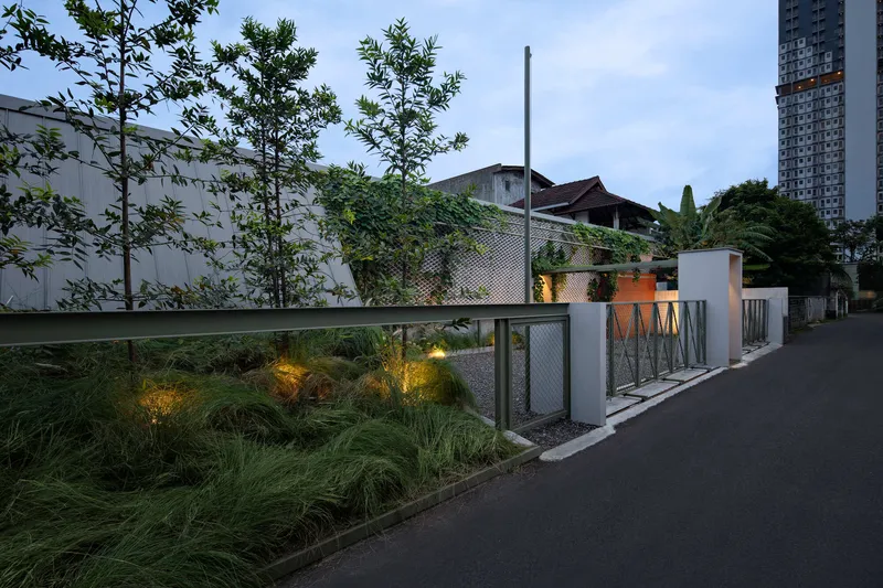 Street view of the perimeter wall with chain-link fencing, vertical slat panels, and planted grasses illuminated at dusk