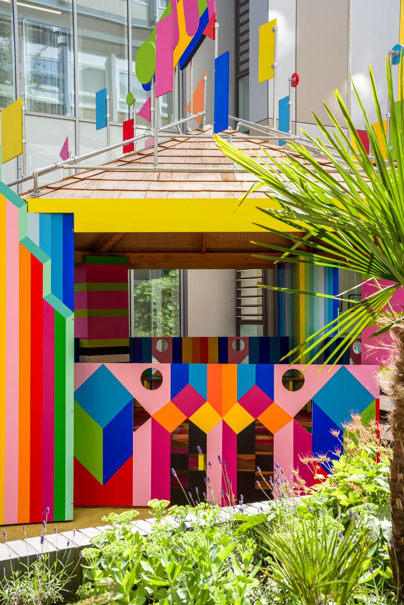 Striped timber roof canopy over multicolored pavilion facade framed by palm fronds and flowering plants