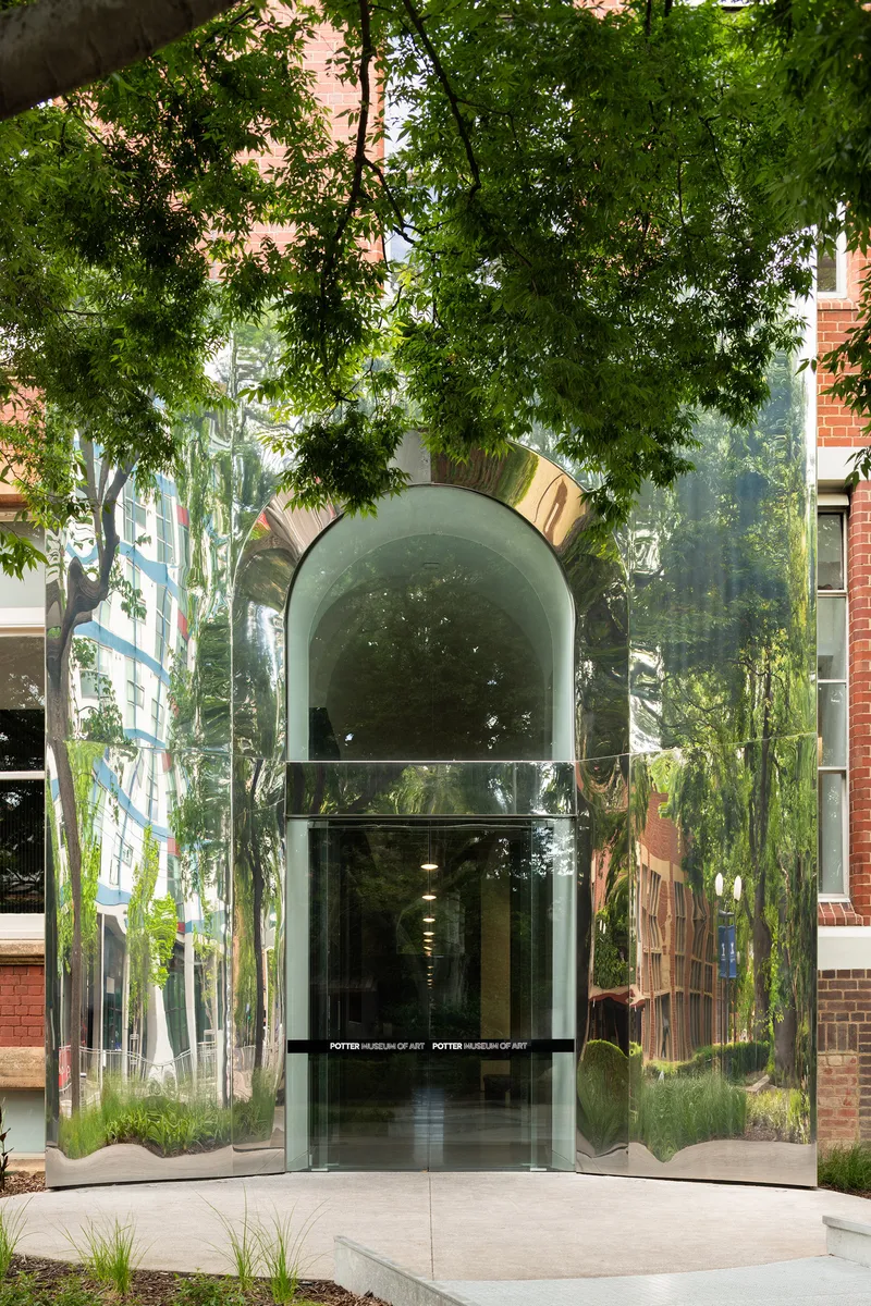 Central glass entrance with mirrored arch canopy beneath overhanging tree branches and dappled shade