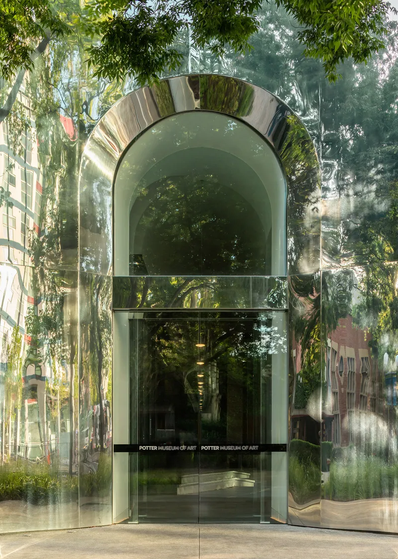 Polished metal arch and glass doorway reflecting foliage and adjacent buildings in afternoon light