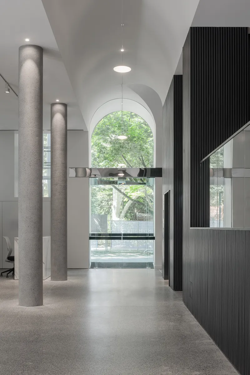 View through vaulted corridor with circular concrete columns toward arched window framing green foliage outside