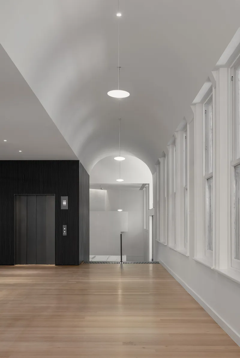 Corridor with vaulted ceiling, pendant lighting, timber floor and vertical black cladding around elevator enclosure