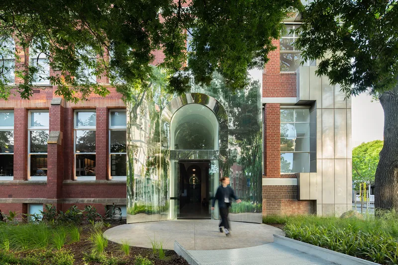 Glass entry pavilion with arched top linking red brick buildings framed by overhanging tree canopy