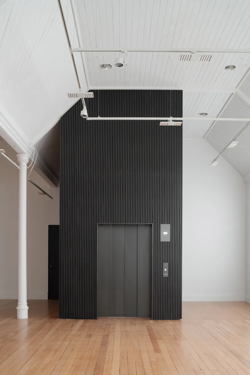 Black ribbed elevator core set against white paneled ceiling and timber floor in gallery hall