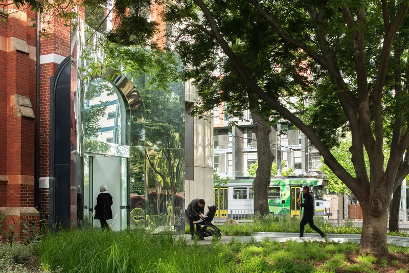Street view of glass pavilion entrance among mature trees with pedestrians and passing tram