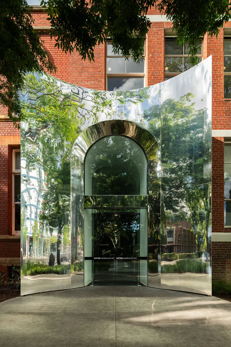 Curved glass entry volume with polished metal arch reflecting surrounding trees and brick walls in daylight