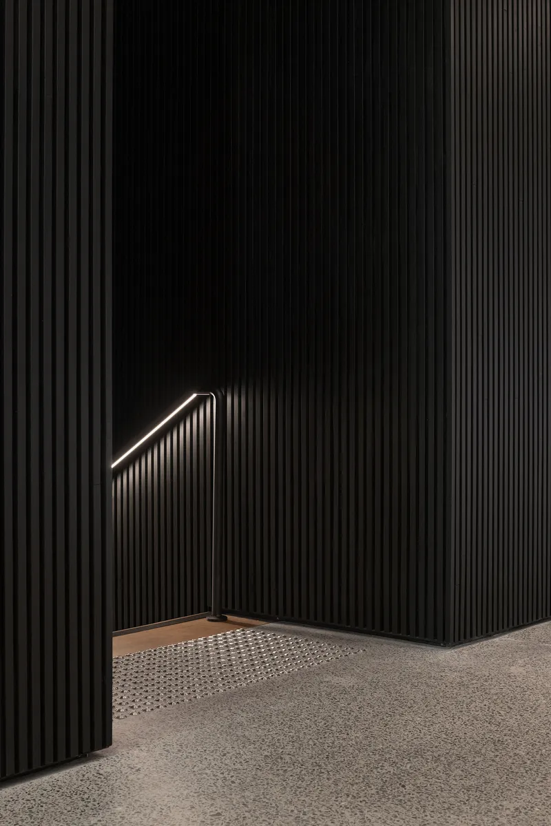 Black vertically-fluted wall with illuminated triangular handrail above a recessed perforated floor panel