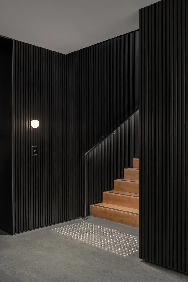 Timber staircase rising between black fluted walls with a circular wall sconce and illuminated floor grille
