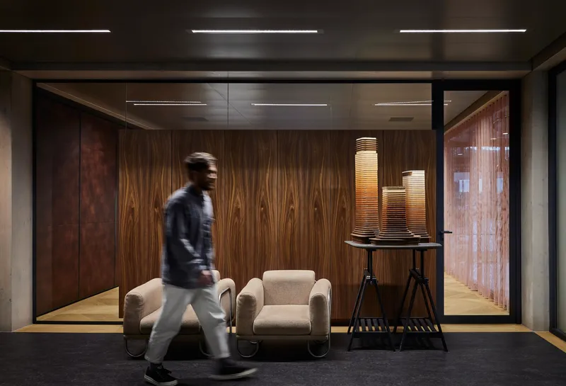 Wood-paneled corridor with glass partition and blurred figure walking past upholstered lounge chairs