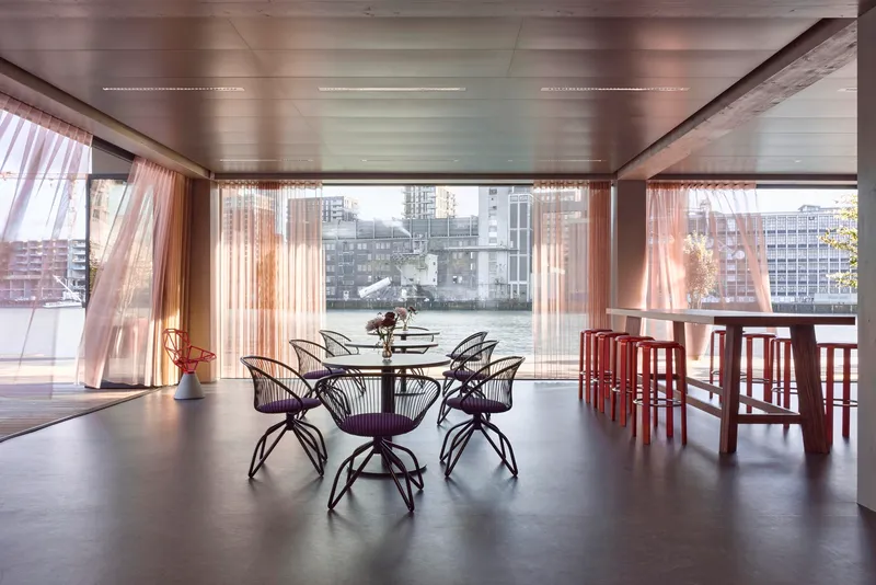 Dining area with metal frame chairs and floor-to-ceiling glazing overlooking a waterfront cityscape