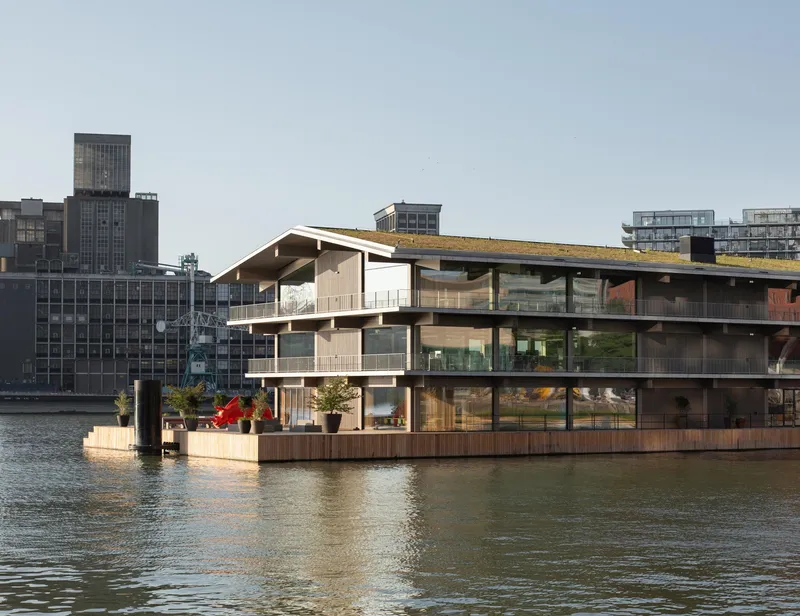 Waterfront view of the three-storey floating structure with timber cladding and green roof under clear skies
