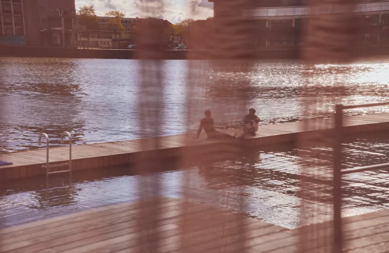 View through sheer curtains to timber swimming dock where two people sit in warm afternoon light