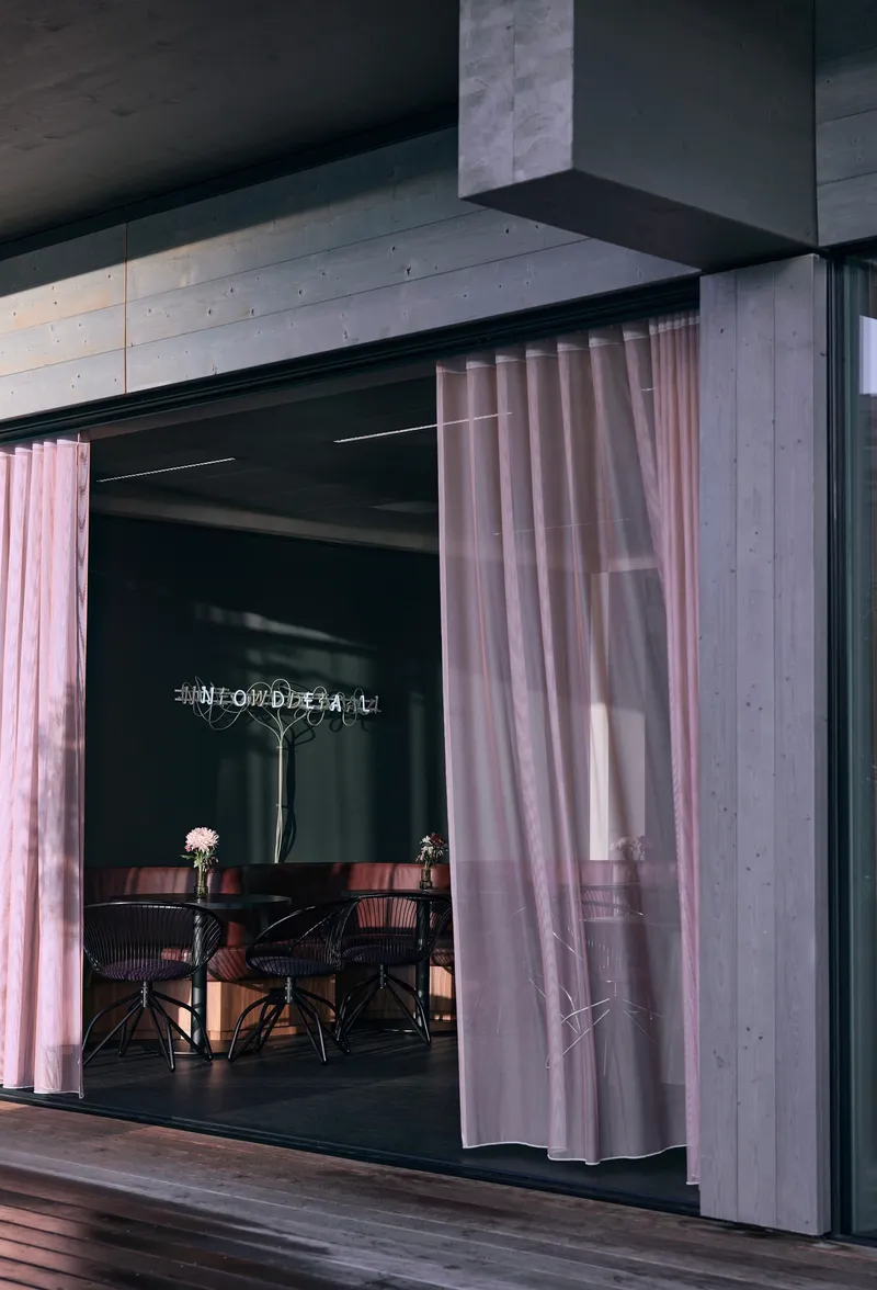 Exterior opening framed by concrete soffit and pink curtains with dining furniture visible inside at dusk