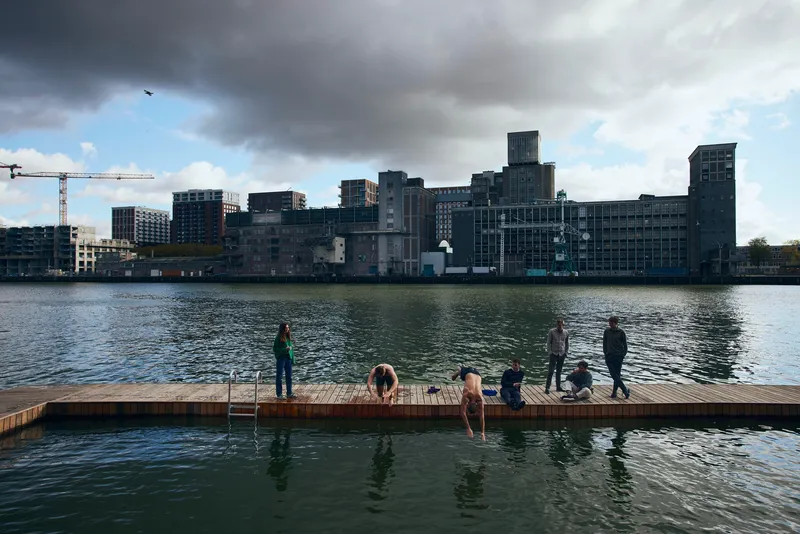 Timber swimming dock with people and a dog facing the converted industrial waterfront under dramatic clouds