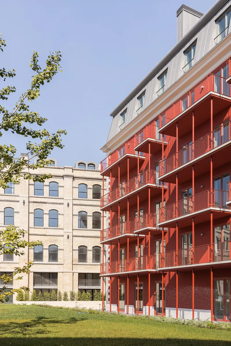 Red metal balconies stacked on a new block facing a historic arched brick building across a lawn