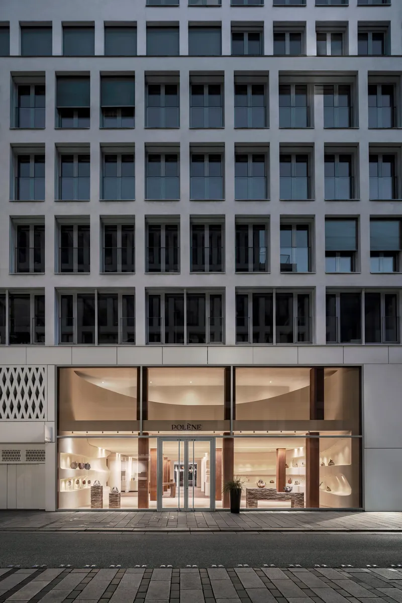Street-level storefront with curved glass facade illuminated at dusk beneath a grey office tower