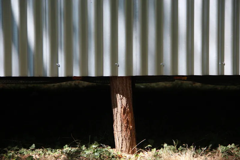 Underside view of corrugated metal wall above timber column base resting on forest floor
