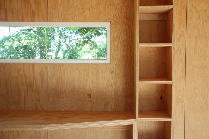 Plywood shelving units and benches beside a horizontal window framing trees in summer foliage