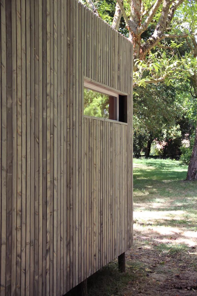 Vertical timber slat facade with single punched window opening among sunlit trees